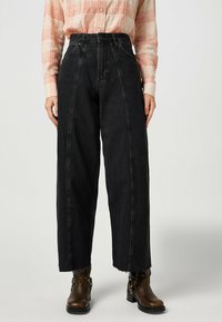 Pantalones de mezclilla negros de cintura alta con pierna ancha, con costuras en contraste y dobladillo deshilachado. Combinados con botines de cuero marrón.