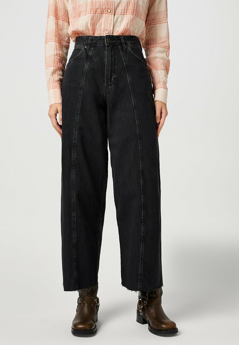 Pantalones de mezclilla negros de cintura alta con pierna ancha, con costuras en contraste y dobladillo deshilachado. Combinados con botines de cuero marrón.