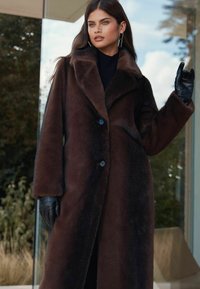 Barna műszőrme kabát széles gallérral, hosszú ujakkal és két fekete gombbal; fekete garbóval és fekete bőrkesztyűvel párosítva.
