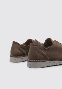 Zapatos de ante marrón con un acabado texturizado, con puntera redonda, detalles cosidos y una suela de goma ligera y moteada. Etiqueta de la marca visible.