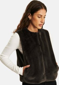 Vrouw met lang, donker haar die gouden oorbellen draagt, een witte langemouwen top en een zwart faux fur mouwloos vest, met haar handen in de zakken.