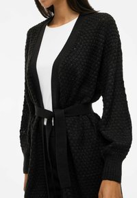 Cardigan noir en maille avec un design texturé, à ouverture frontale et manches longues. Doté d'une ceinture en tissu assorti à la taille pour un ajustement.