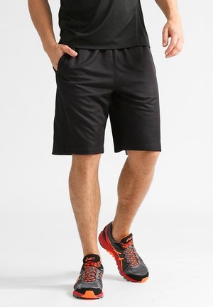 Mand iført sorte atletikshorts og sorte og orange løbesko, stående med den ene hånd i lommen mod hvid baggrund.
