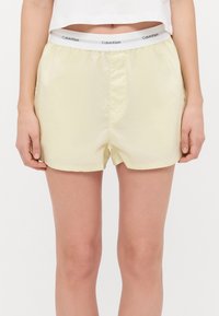 Lichtgele shorts gemaakt van gladde stof met een elastische tailleband waarop het Calvin Klein-logo in het grijs staat. De zoom eindigt halverwege de dij.
