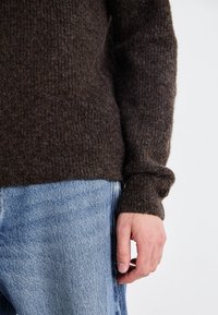 Pull en tricot marron avec un motif texturé, doté de poignets côtelés et d'une coupe décontractée, associé à un jean en denim bleu clair.