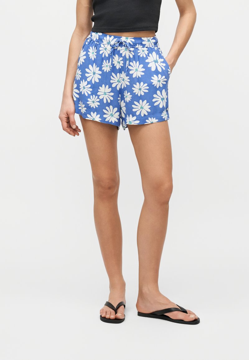 Γυναίκα που φοράει μπλε σορτς με λευκό τύπωμα μαργαρίτας, μαύρο crop top και μαύρα σαγιονάρες, στέκεται με το ένα χέρι στην τσέπη.