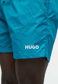 Κοντινό ενός ατόμου που φοράει φωτεινά τυρκουάζ σορτς με το λογότυπο "HUGO" τυπωμένο σε λευκό κοντά στο τελείωμα, σε σκουρόχρωμο δέρμα.