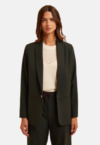 Blazer verde scuro con un bottone singolo, due tasche frontali e una vestibilità sartoriale, abbinato a un top crema chiaro con un motivo decorativo.