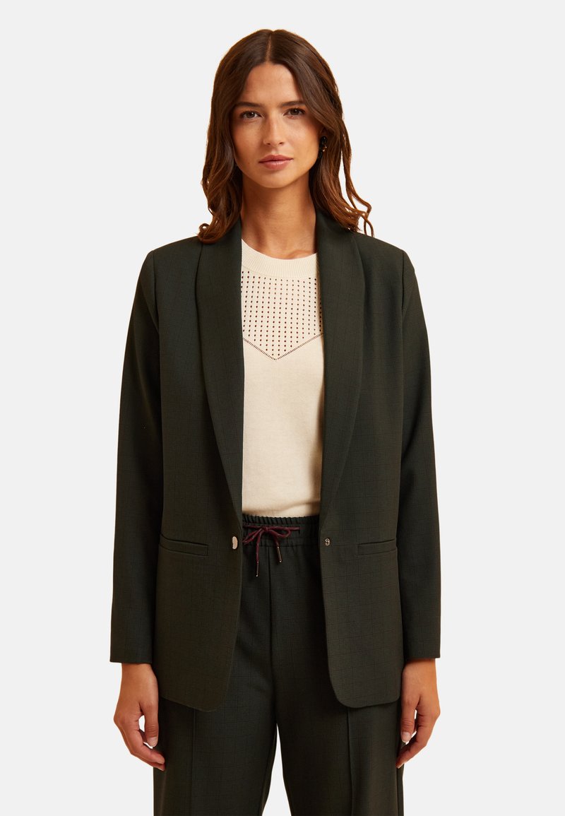 Blazer verde scuro con un bottone singolo, due tasche frontali e una vestibilità sartoriale, abbinato a un top crema chiaro con un motivo decorativo.