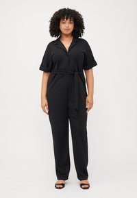 Curly-haired woman stands confidently in a black belted jumpsuit, minimalist style, with a clean white background.