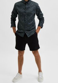 Tmavě modrá košile s knoflíky a malým květinovým vzorem, v kombinaci s černými šortkami a světle šedými teniskami. Košile má dlouhé rukávy a límec.