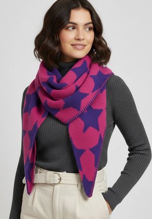 Woman wearing a pink and purple star-patterned scarf over a dark gray ribbed sweater and light beige high-waisted pants with a belt.