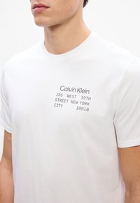 Tricou alb din bumbac cu text gri tipărit, care conține numele brandului și detalii despre adresă. Mâneci scurte și guler clasic tip crew.