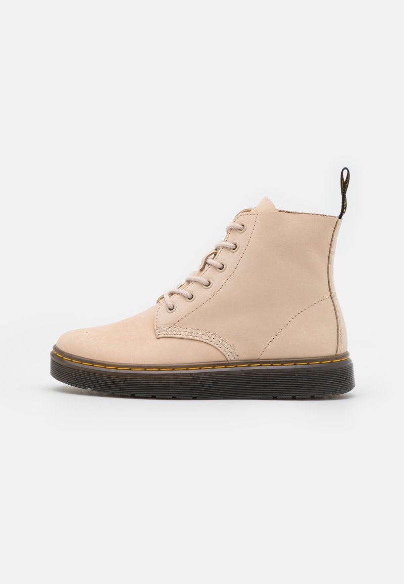 Bottine en cuir beige à lacets, semelle noire et coutures jaunes, avec une languette à l’arrière.