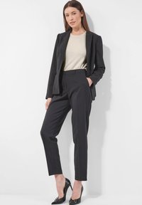 Frau in schwarzem Blazer und Hosen mit beigem Oberteil und schwarzen High Heels, die vor einem weißen Hintergrund mit der Hand in der Tasche steht.