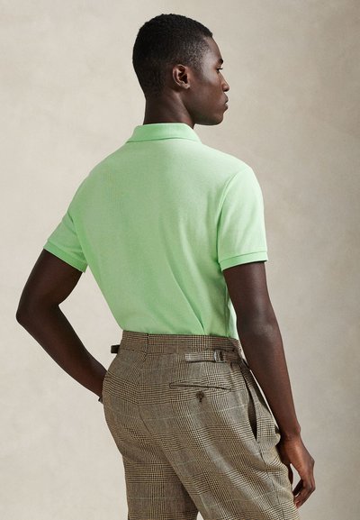 Πράσινο πόλο με κοντά μανίκια, σε συνδυασμό με καφέ καρό παντελόνι. Η υφή του πουκαμίσου είναι λεία και ελαφριά, διαθέτοντας κλασικό γιακά.