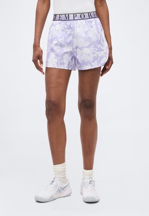 Frau trägt lila-weiß gemusterte Shorts mit elastischem Bund und der Aufschrift "Emporio", weiße Socken und weiße Sportschuhe.