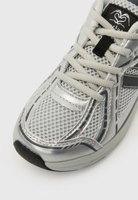 Sølvfarget atletisk sko med mesh- og syntetisk overdel. Reflekterende detaljer, lysegrå lisser og svarte aksenter på sålen og merket.