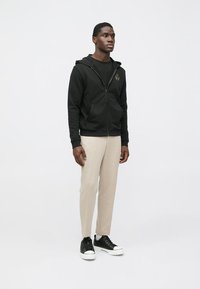 Juodas užtraukiamas džemperis su kišenėmis, derinamas su šviesiai smėlio spalvos kelnėmis. Modelis dėvi juodus sportbačius su baltais padais. Paprastas dizainas, be matomų raštų.