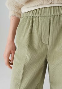Helle grüne Cordhose mit strukturiertem Finish, ausgestattet mit einem elastischen Bund und Detail-Taschen auf der Vorderseite. Sichtbare Hand auf dem Bund.