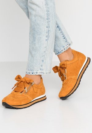 Persoon met lichtgewassen jeans en oranje suède sneakers met dikke veters, zijritsen en witte zolen, staand op een lichte vloer.