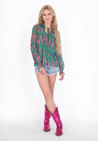 Blusa a maniche lunghe verde e rosa a fantasia con chiusura a bottone, abbinata a pantaloni shorts in denim strappati e stivali da cowboy rosa lucidi.