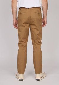 Homme portant un pantalon marron et des baskets blanches, se tenant avec les mains relaxées sur les côtés, vu de dos sur un fond uni.