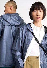Des vestes de pluie légères et bleues avec capuches, dotées de boutons-pression et de cordons réglables, portées par deux personnes faisant face dans des directions opposées.