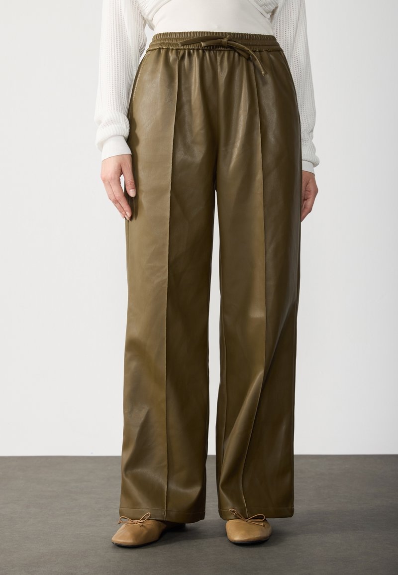 Personne portant un pantalon large en similicuir vert olive avec taille à cordon, un pull en maille blanc et des ballerines beige, debout sur un sol gris.