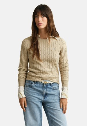 Frau mit langen braunen Haaren, die einen beige gestrickten Zopfpullover über einem weißen Hemd mit Muster und hellblauen hoch taillierten Jeans trägt, steht vor einem weißen Hintergrund.