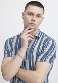 Camicia a maniche corte con righe blu, verdi e bianche. Caratterizzata da un colletto con bottoni e una trama sottile. Modello con la mano appoggiata sul mento.