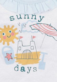 Valkoisessa kankaassa on värikäs kuva, jossa on keltainen aurinko, rantalinna, tähtiä ja sateenvarjo, jonka vieressä on teksti "aurinkoiset päivät".