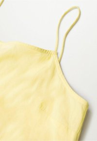 Gros plan sur un haut sans manches jaune clair avec de fines bretelles spaghetti sur fond blanc.