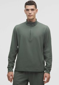 Tmavozelený športový top s dlhým rukávom, s elastickou golierom so zipsom a textúrovaným vzorom. Hladký povrch a uvoľnený strih.