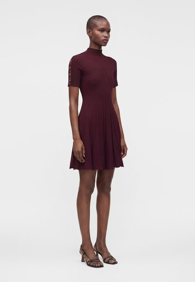 Femme portant une robe courte bordeaux côtelée à manches courtes avec des boutons sur les manches et des sandales noires à talons avec brides.