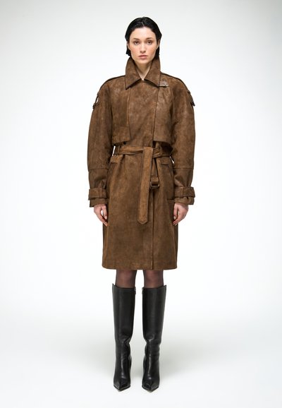 Žena v hnedom semišovom trenčkotom so zónou v páse, prackami na manžetách a čiernymi čižmami nad kolená stojí na bielom pozadí.