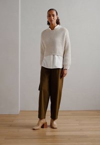 Beige kort tröja med ribbad textur över en vit skjorta, i kombination med olivgröna byxor och sandfärgade klackkängor.