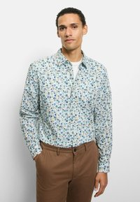 Hellblaues Hemd mit Knöpfen und floralem Muster in Grün, Blau und Beige. Lange Ärmel und ein klassischer Kragen. Kombiniert mit braunen Hosen.