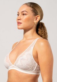Valkoinen pitsinen bralette, jossa on läpikuultava muotoilu, kolmiomaiset kupit, ohuet hihnat ja aallotettu reuna. Sileä kangas hienoilla yksityiskohdilla.