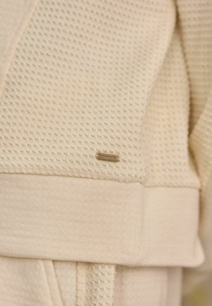 Beige textúrájú anyag vaffle mintával, sima mandzsettával és egy kis arany "DOWNLINE" címkével. Részletes varrás látható.