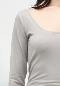 Γυναίκα που φοράει ανοιχτό γκρι πουκάμισο με μακριά μανίκια και στρογγυλή λαιμόκοψη, εμφανιζόμενη από τον λαιμό έως τη μέση σε λευκό φόντο.