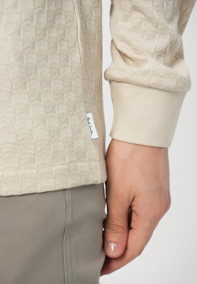 Μπεζ μακρυμάνικο πουκάμισο με υφή και υφαντό σχέδιο, με ριμπ μανσέτες και μικρή ετικέτα στο τελείωμα. Συνδυασμένο με γκρι παντελόνι.