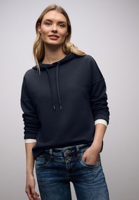 Jeune femme portant un sweat à capuche noir texturé par-dessus une chemise blanche et un jean en denim bleu, une main dans la poche, sur un fond gris clair.