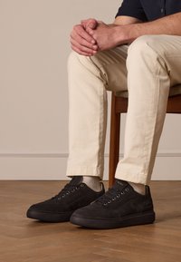 Baskets basses en daim noir avec une semelle en caoutchouc, dotées d'œillets dorés et d'un design épuré. Portées avec un pantalon couleur crème et des chaussettes légères.