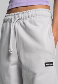 Hellgraue Jogginghosen aus weichem Material, ausgestattet mit einem elastischen Taillenbund und Kordeln sowie einem schwarzen Logo-Patch an der Seite.