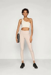 Top crop bianco sporco con una spalla, abbinato a leggings rosa chiaro e scarpe da ginnastica nere, mentre tiene una borraccia nera, in piedi contro uno sfondo grigio.