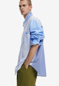 Hellblau-weißen gestreiften Button-Up-Hemd mit lockerem Schnitt, mit einer Tasche und hochgekrempelten Ärmeln. Kombiniert mit grünen Hosen.