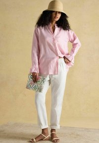 Cămașă roz cu dungi și nasturi, cu mansete cu volane, pantaloni albi lejeri și sandale plate. Ține o geantă plic cu model floral.