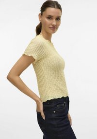 Lys gul, teksturert kortermet topp med en bølget halskant og kant, kombinert med mørke denimjeans.