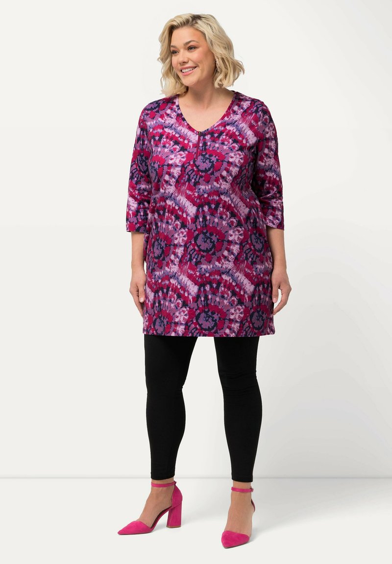 Vibrant lilla tunika med blomstermønster, ¾ ærmer og V-hals, parret med sorte leggings og lyserøde hæle.